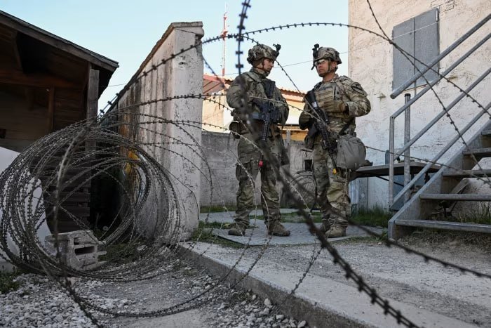 Two soldiers in camouflage uniforms and helmets, carrying rifles, stand conversing behind coils of concertina wire near a concrete building and metal staircase.