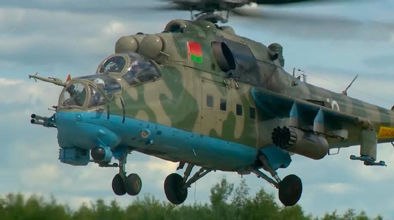 A camouflage-painted military helicopter with a blue underside hovers low over a green treeline under a cloudy sky.