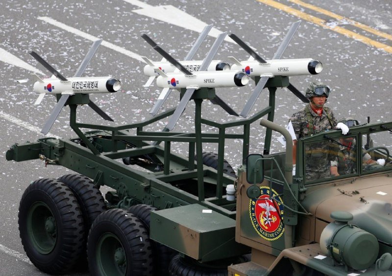 A military parade features a camouflaged truck carrying mounted white "SPIKE" missiles, with uniformed personnel in helmets riding in the vehicle.