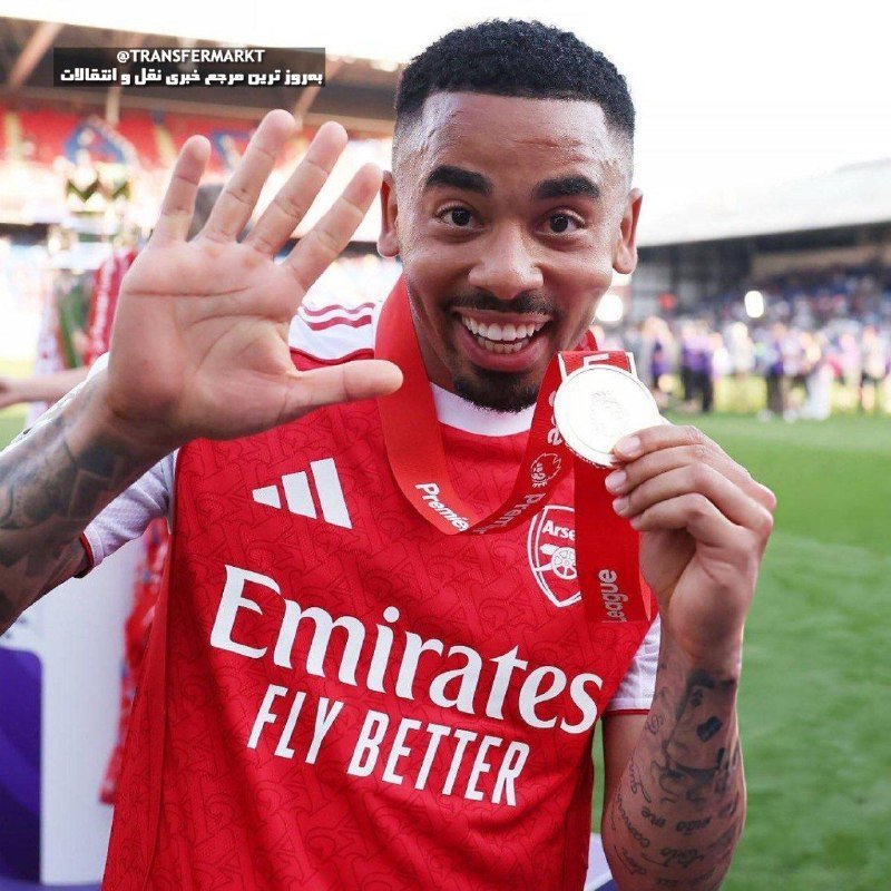 A smiling man in a red Arsenal jersey with "Emirates Fly Better" sponsorship holds up a silver medal on a football pitch.