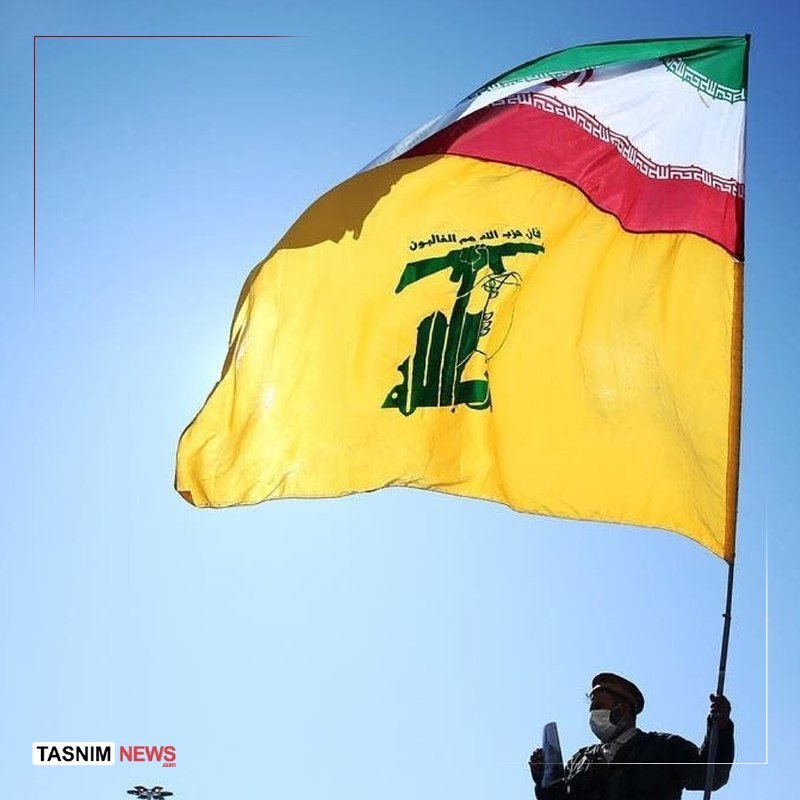 A masked person in uniform waves a large yellow Hezbollah flag against a clear blue sky.