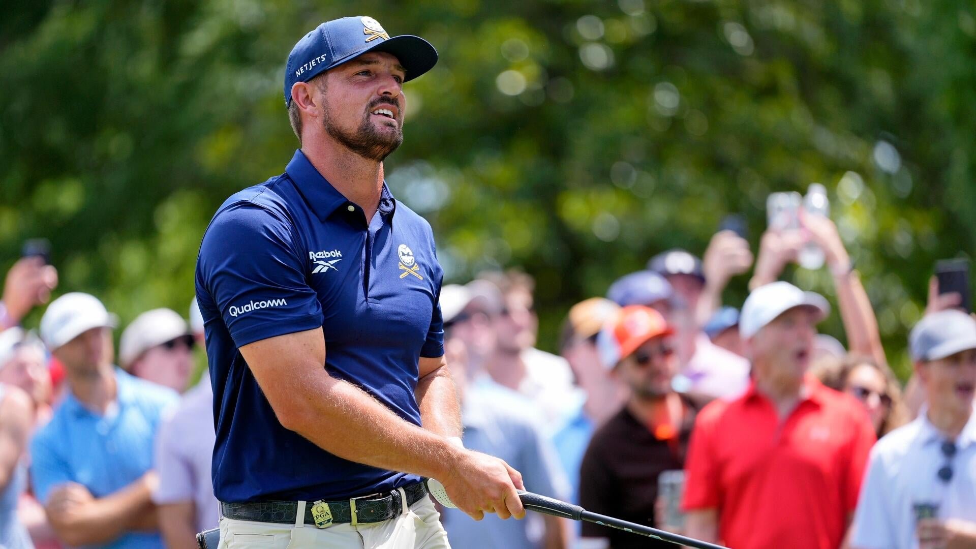 A bearded golfer in a navy Reebok polo and "NETJETS" cap watches a shot mid-swing, surrounded by a crowd of spectators under trees.