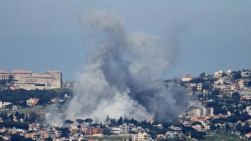 A large plume of gray smoke rises above a hillside community with densely packed buildings under a hazy sky.