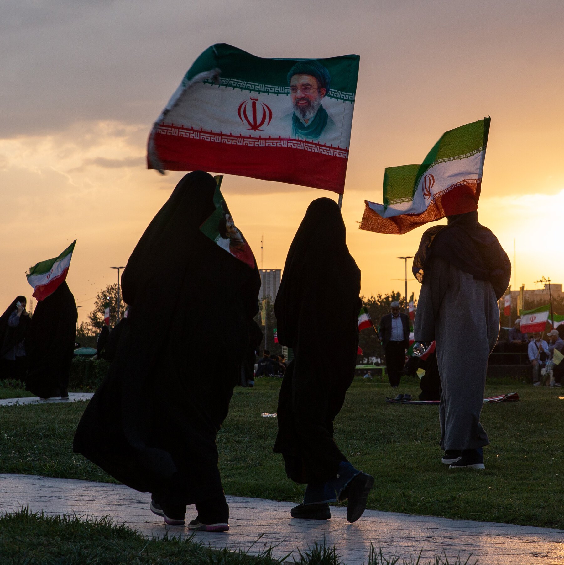 People in black chadors and a gray cloak walk along a paved path carrying Iranian flags, one bearing a portrait of a bearded man in a turban, at sunset.