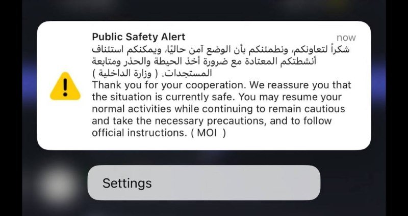 A smartphone screen displays a "Public Safety Alert" notification in English and Arabic with a yellow warning icon, reassuring recipients the situation is safe and advising them to follow official instructions.