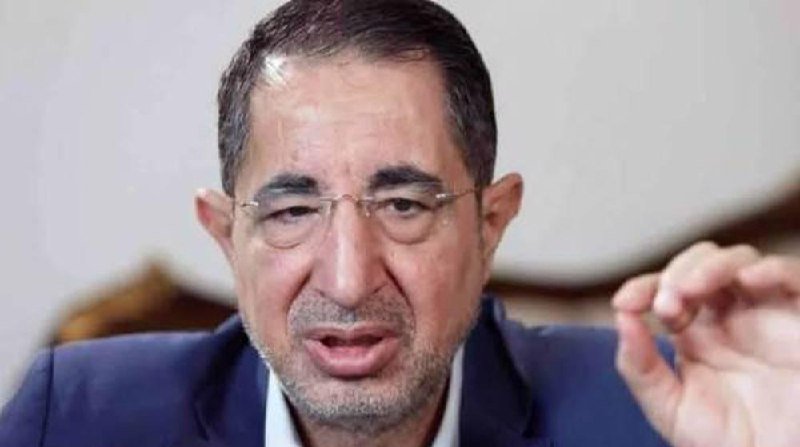 A man in a dark suit and glasses speaks while gesturing with one hand raised, photographed in a close-up shot against a blurred light background.