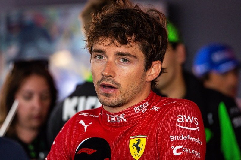 A curly-haired man in a red Ferrari racing suit with sponsor logos speaks into a microphone during an interview.