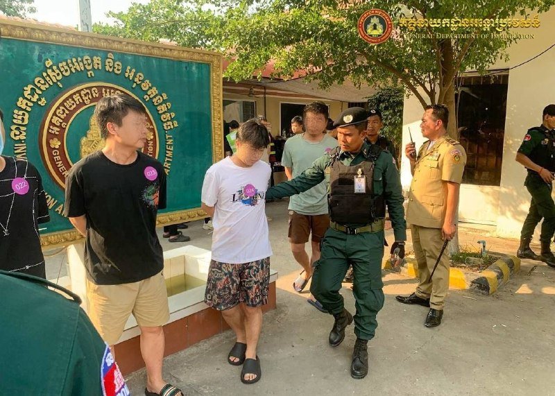 Uniformed officers escort handcuffed individuals past a green government department sign outdoors.