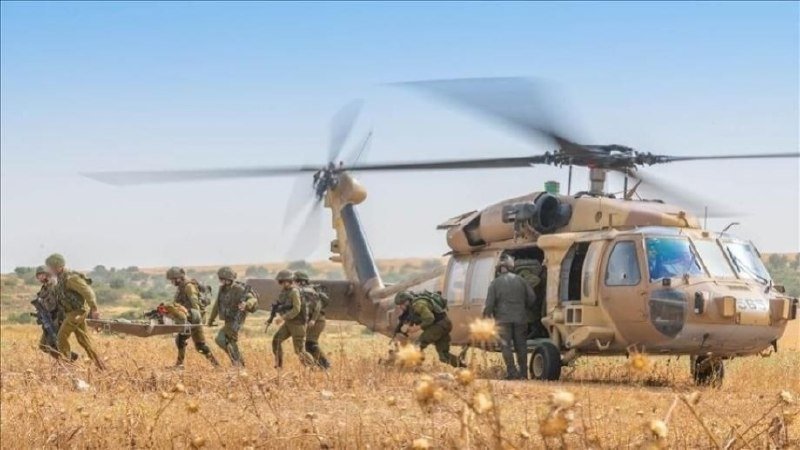 Soldiers in green uniforms carrying a stretcher run across a dry field toward a tan military helicopter with spinning rotor blades under a clear blue sky.