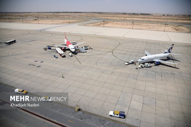 Two commercial airplanes are parked at an airport tarmac surrounded by ground service vehicles, with a desert landscape visible in the background.