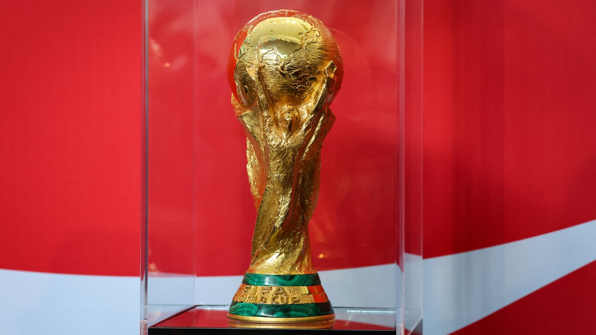 A gold FIFA World Cup trophy sits inside a clear display case against a red and white background.