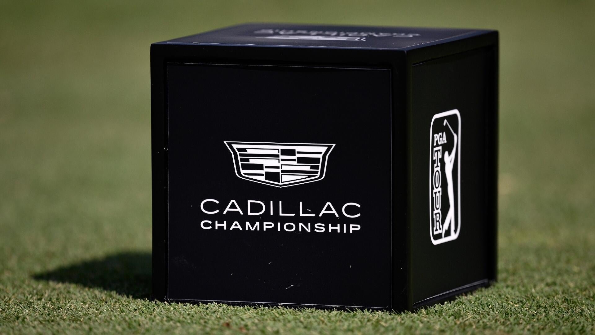 A black cubic tee marker displaying the Cadillac Championship logo and PGA Tour logo sits on a grassy golf course.