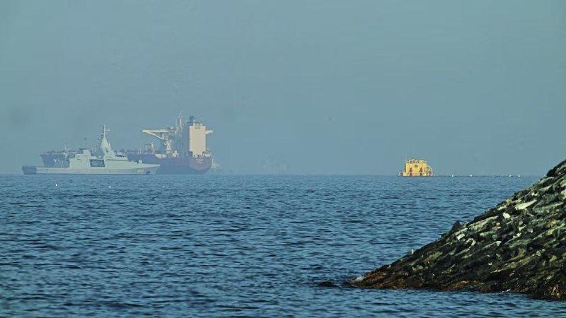 A naval vessel and a cargo ship sit on calm, hazy waters offshore, with a rocky shoreline visible in the foreground.