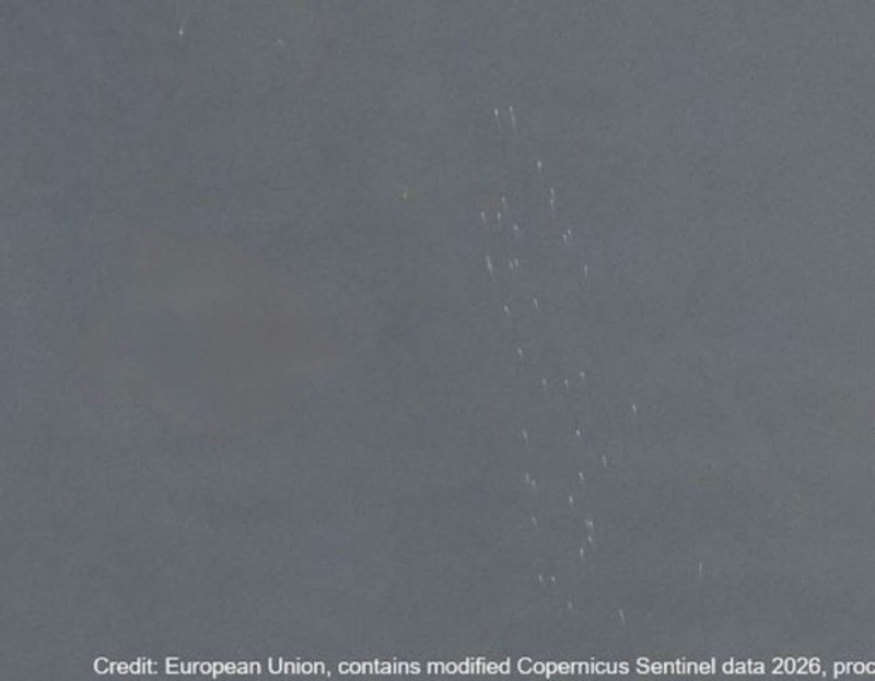 A satellite image shows a gray-blue ocean surface with a diagonal trail of small white objects and a dark patch visible in the water.
