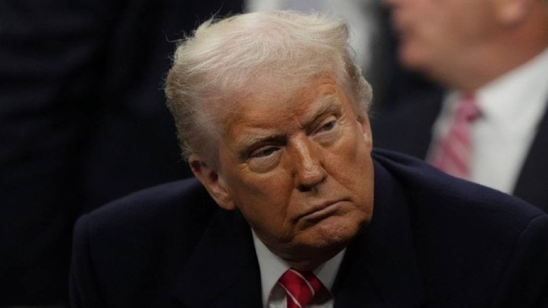 A man with light blonde hair wearing a dark suit and red striped tie looks downward, with another suited figure partially visible behind him.