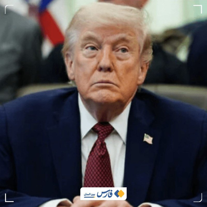 An older man with blonde hair wearing a dark suit, white shirt, and red patterned tie with an American flag pin sits in front of a blurred American flag.