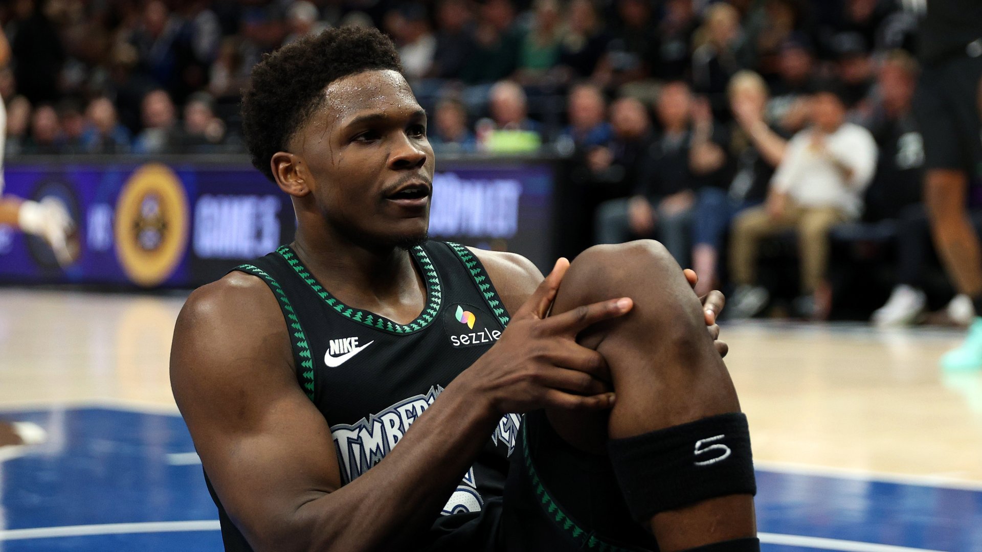 A basketball player in a black Timberwolves jersey with "Sezzle" and Nike logos sits on a court holding his knee, with a blurred crowd in the background.
