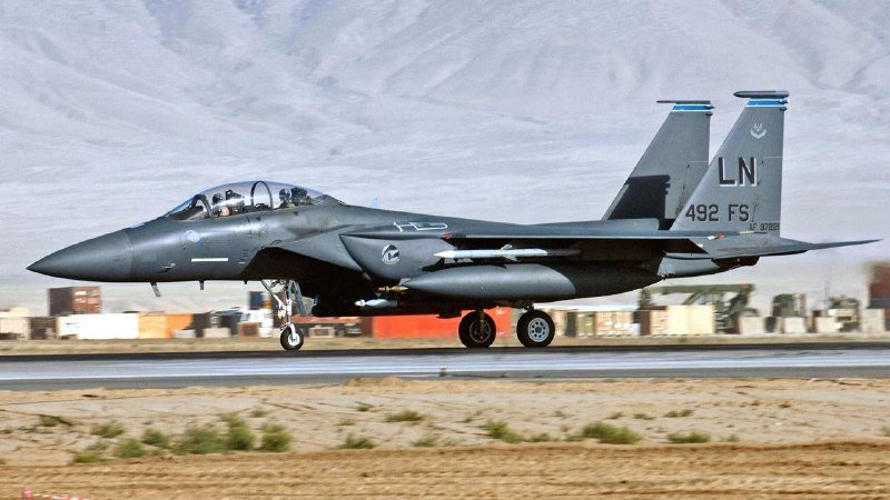 A gray military fighter jet marked "LN" and "492 FS" lands on a runway with two pilots visible in the cockpit, set against a mountainous desert backdrop.