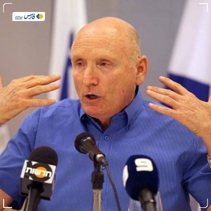 A bald, older man in a blue striped shirt speaks at a podium with microphones, gesturing with both hands raised, in front of a blue and white flag.