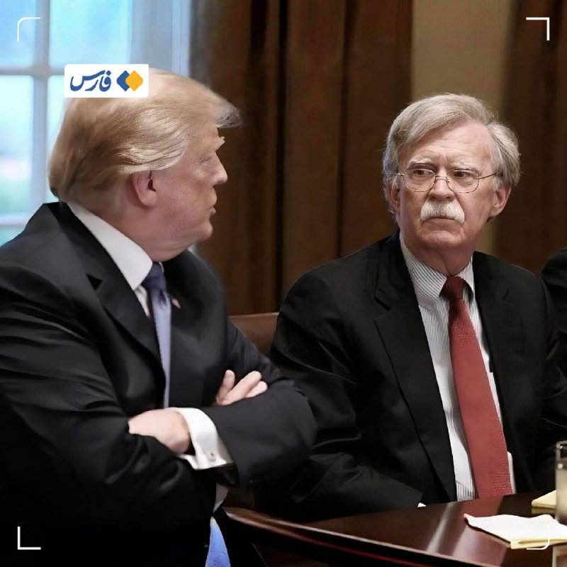 Two men in suits sit at a wooden table in a formal room with curtains, one with blonde hair and crossed arms, the other wearing glasses and a red tie.