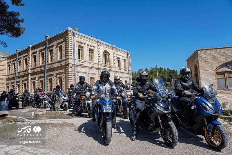 A group of helmeted motorcyclists on sport and touring bikes gather outside a historic brick building under a clear blue sky, with a "Fars News" watermark visible.