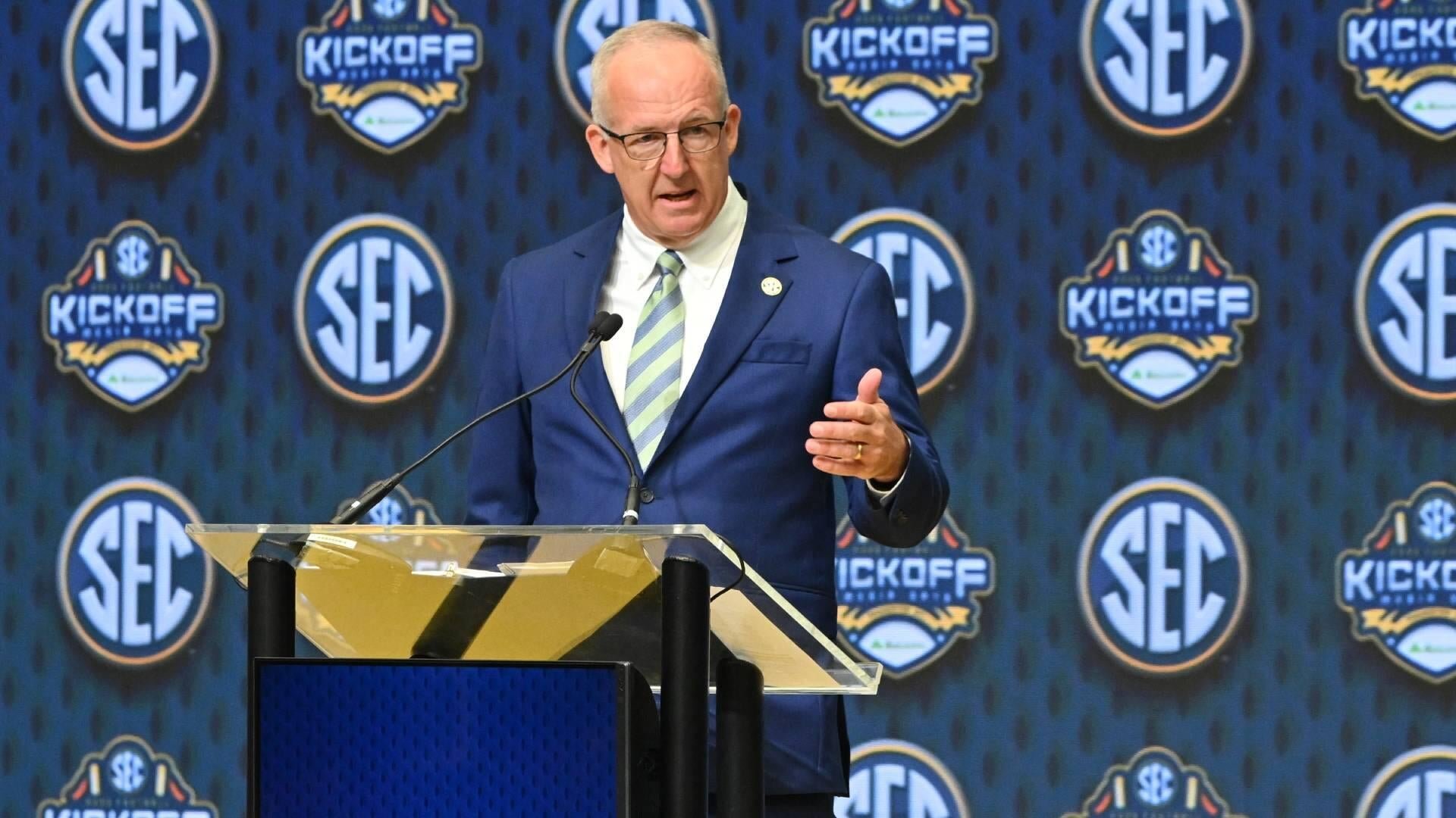 A man in a blue suit and glasses speaks at a clear podium, gesturing with his hand, in front of a backdrop displaying SEC and SEC Kickoff logos.