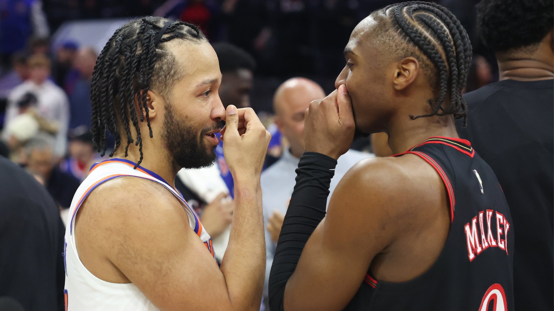 Two basketball players in opposing jerseys—one white with orange and blue trim, one black with red trim—stand face-to-face in a crowded arena, appearing to exchange words.