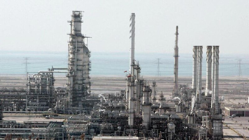 A large industrial refinery or petrochemical plant with tall smokestacks and complex piping stretches across a flat coastal landscape under a hazy sky.