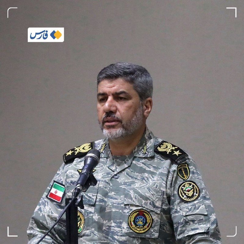 A man with a gray beard in a camouflage military uniform stands at a microphone, with an Iranian flag patch visible on his sleeve.