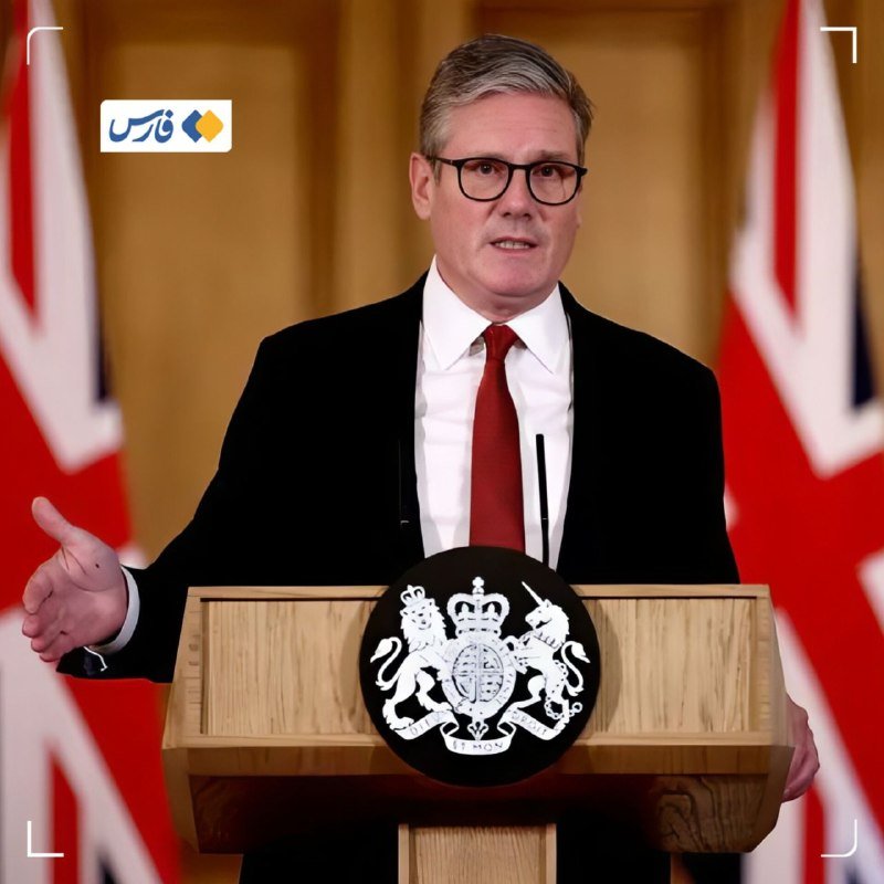 A man in a dark suit and red tie stands at a wooden lectern bearing a royal crest, gesturing with one hand in front of British Union Jack flags.