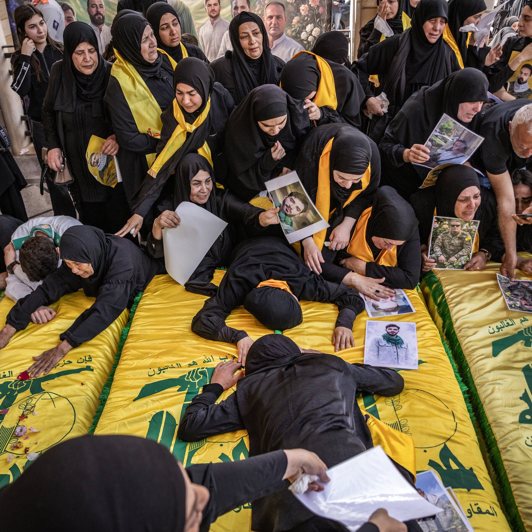 Women in black mourning attire and yellow sashes gather around yellow-draped coffins, holding photographs of young men in a crowded funeral ceremony.