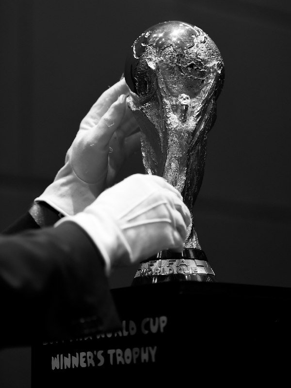 White-gloved hands carefully handle a golden FIFA World Cup trophy displayed on a black pedestal labeled "WINNER'S TROPHY" in this black-and-white photograph.