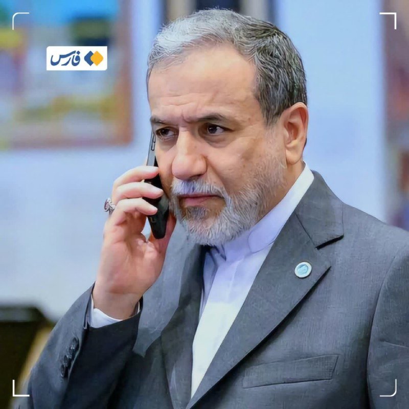 A gray-haired, bearded man in a dark suit and light blue shirt speaks on a black smartphone, with a Persian-script logo visible in the corner.