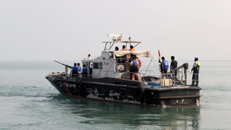 A patrol boat labeled "PILOT" floats on calm waters with armed personnel and crew in life vests standing on deck.