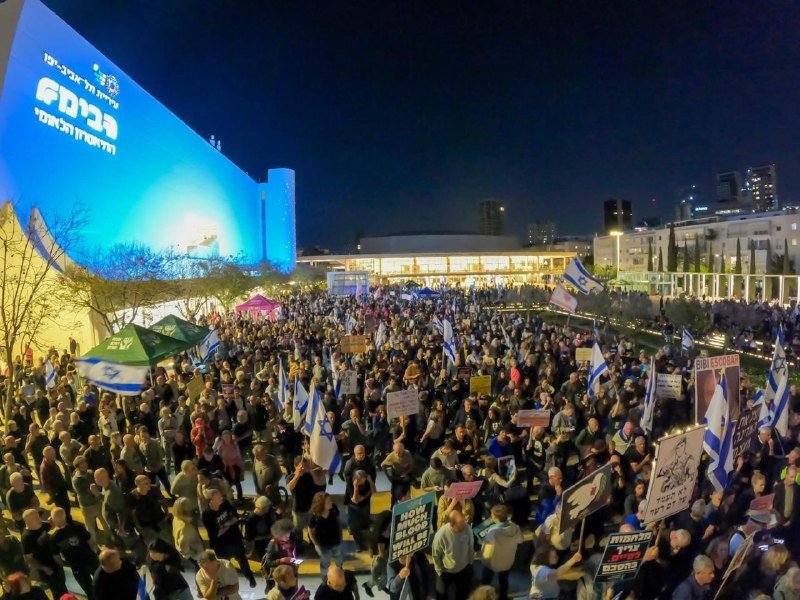 A large crowd gathers at night outside an illuminated building displaying a blue banner with Hebrew text, holding Israeli flags and protest signs.