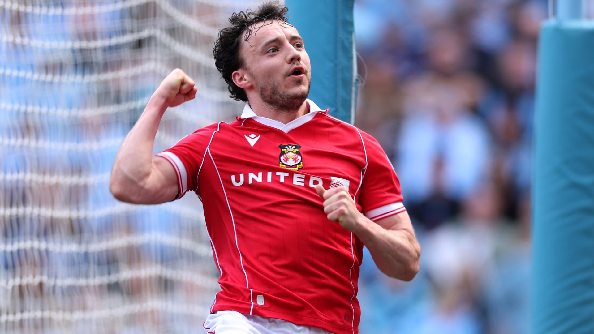 A soccer player in a red "UNITED" jersey clenches his fist and pumps his arms in celebration near the goal netting, with blurred spectators in the background.