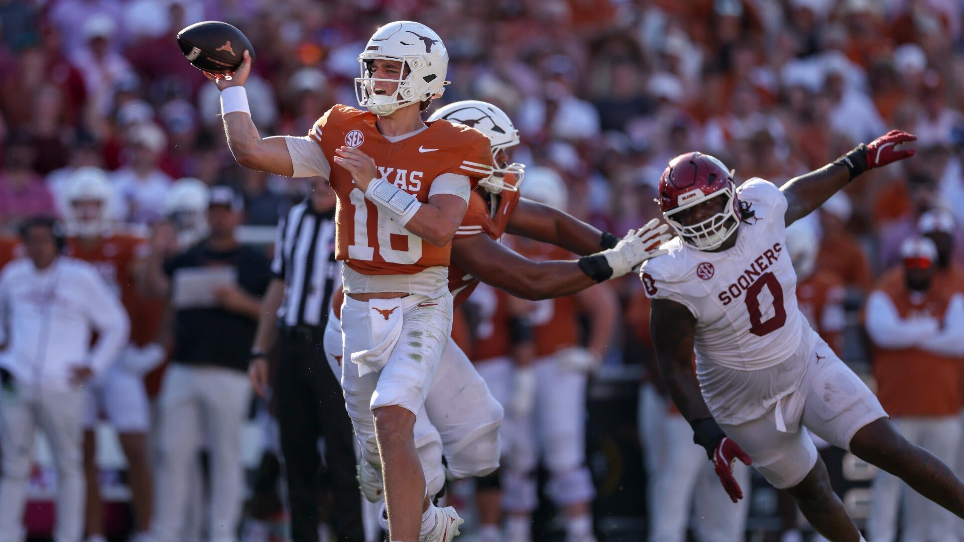 A Texas quarterback in an orange jersey throws a football while being pressured by a Sooners defender in a white and crimson uniform during a college football game.