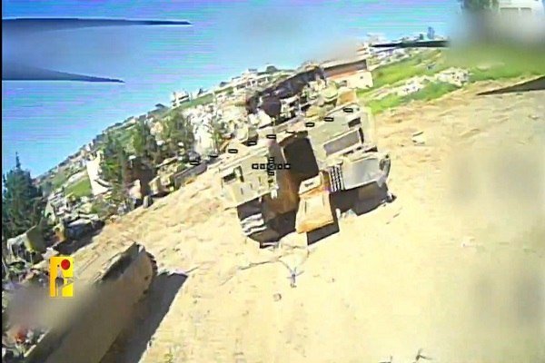 A camouflaged military vehicle drives along a sandy, war-damaged street near waterfront buildings in the background.