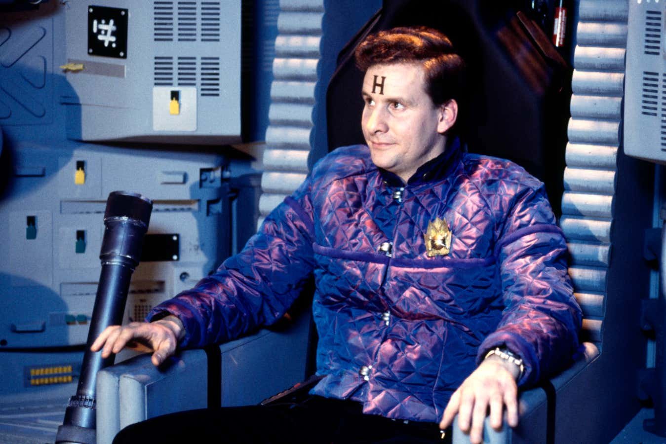 Actor in a purple quilted jacket with an "H" on his forehead sits in a sci-fi set chair, surrounded by metallic wall panels.