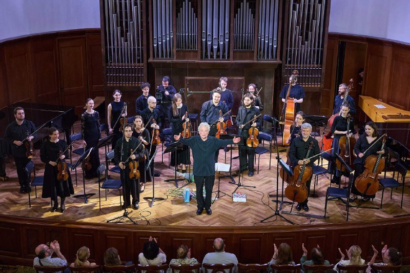 A conductor in dark clothing stands center stage before a string ensemble holding their instruments on a wooden concert hall platform, as audience members in the foreground applaud.