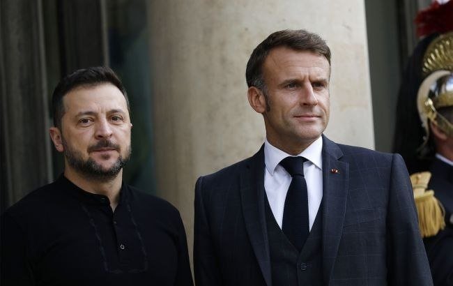 Two men stand side by side, one in a dark suit with a tie and lapel pin, the other in a black collared shirt, beside a uniformed guard with a plumed helmet.
