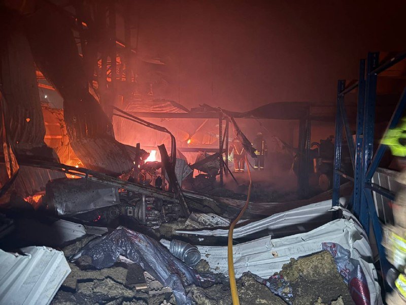 Firefighters stand amid smoke-filled, debris-strewn wreckage of a destroyed structure with visible flames and an orange glow.