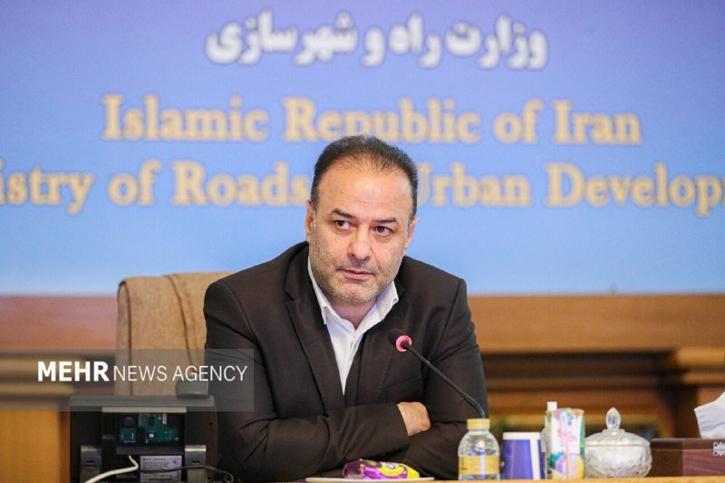 A man in a dark suit sits at a desk with a microphone, speaking in front of a backdrop reading "Islamic Republic of Iran" and "Ministry of Roads and Urban Development."