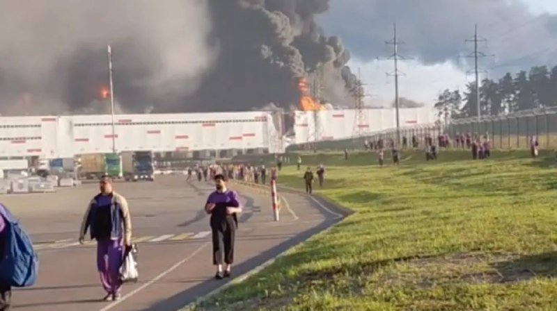 Employees walk away from a large industrial building with visible flames and thick black smoke rising from its roof.
