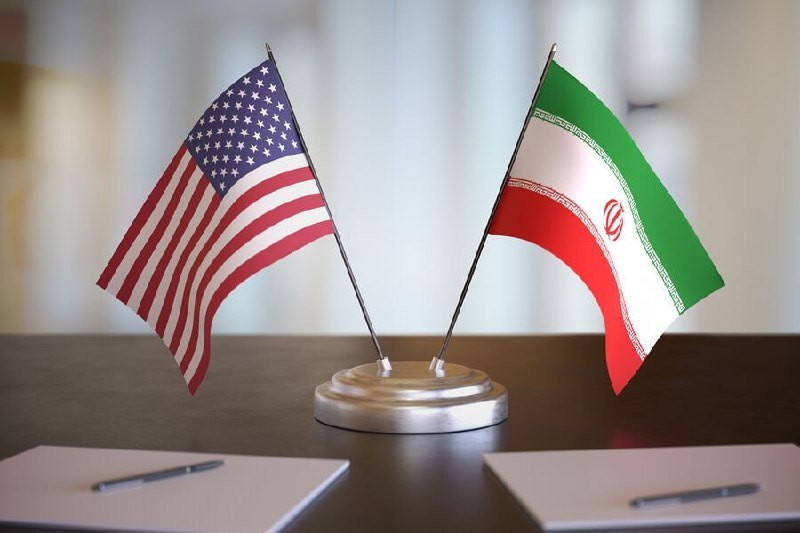 Two small desk flags of the United States and Iran stand crossed on a silver base, with two blank white papers and pens placed on a dark table.