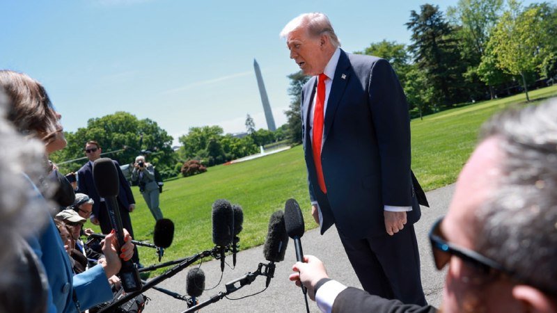 An older man in a dark suit and red tie speaks to reporters holding microphones, with the Washington Monument visible in the background on a sunny day.