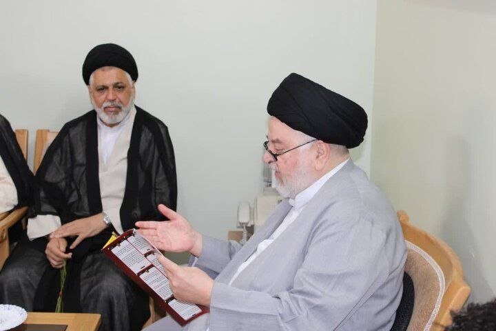 Two men wearing black turbans and clerical robes sit indoors, one gesturing while holding an open notebook and pen.