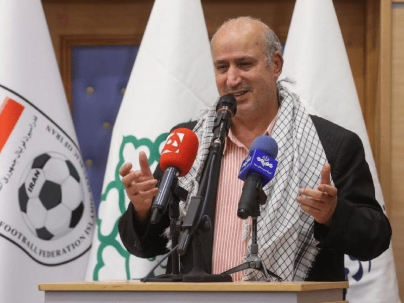 A smiling man wearing a checkered scarf speaks at a podium with multiple microphones, with the Iran Football Federation logo visible on a flag behind him.