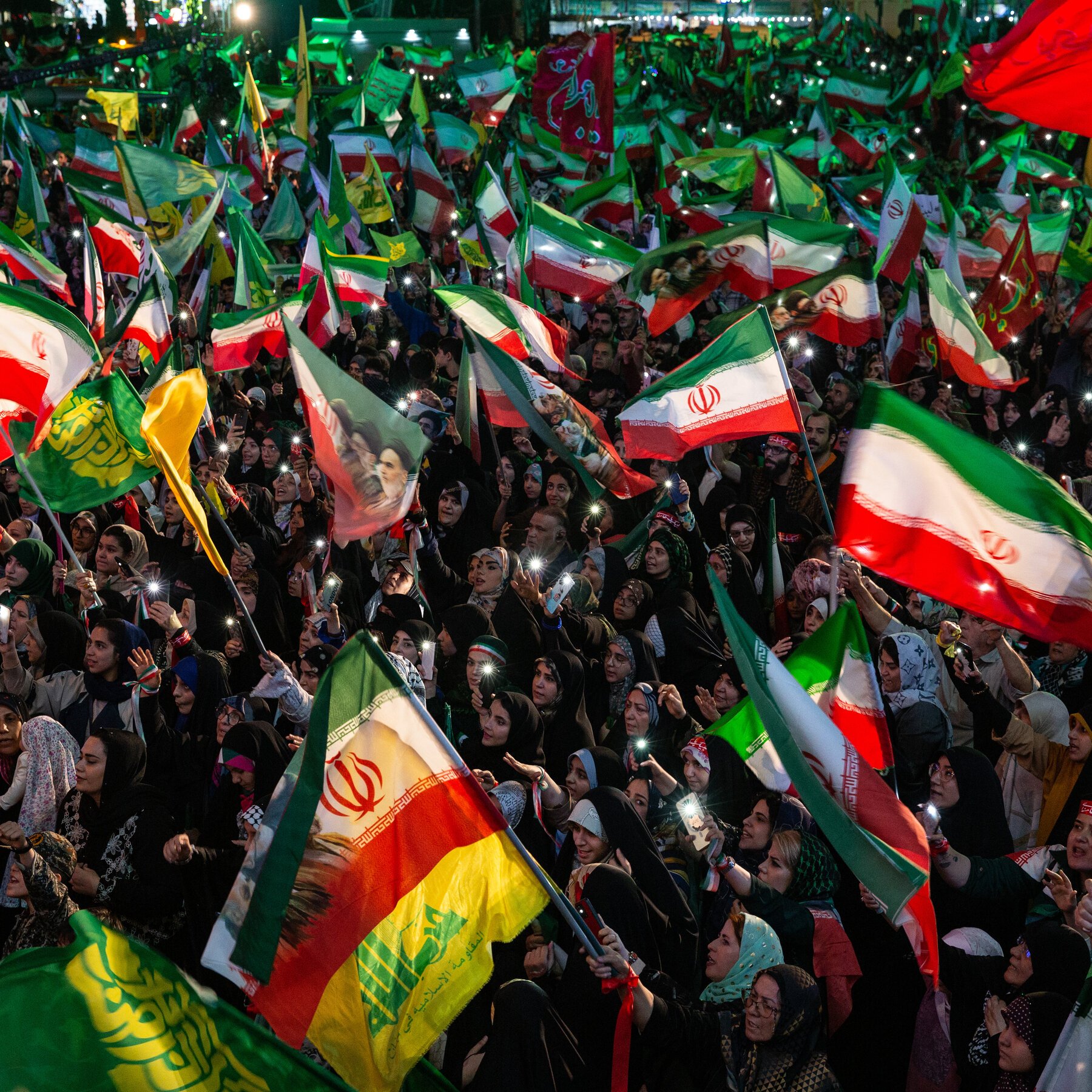 A large nighttime crowd waves Iranian flags and illuminated phone lights while cheering during a public rally.