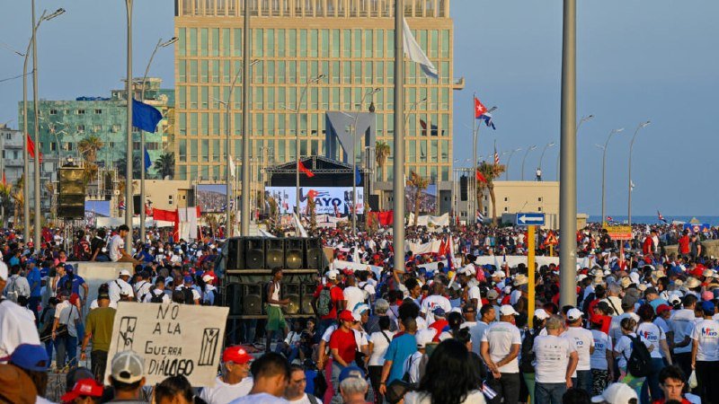 A large crowd gathers in an outdoor public square holding flags and a sign reading "NO A LA GUERRA DILMAED" before a tall building and stage.